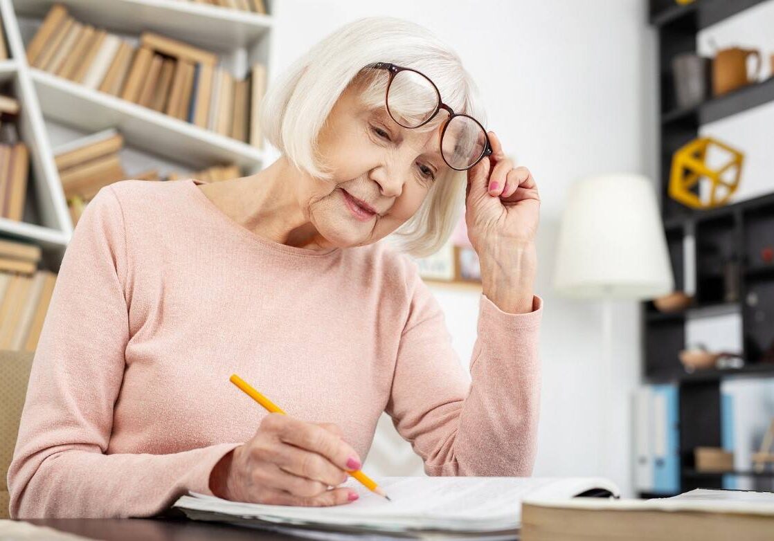 Elderly woman writing in a notebook while adjusting her glasses