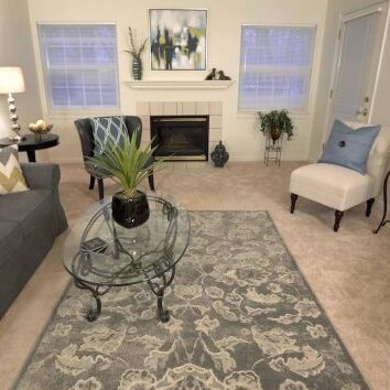 A spacious living room in an independent senior living residential apartment at Silver Maples. The inviting space includes a fireplace, sofa, armchairs, coffee table, artwork, rug, plants, and windows.