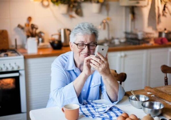 Senior woman using mobile phone at cozy home kitchen. Retired person shopping online. People connection, communication with gadget, delivery in isolation, distance healthcare. Lifestyle moment.