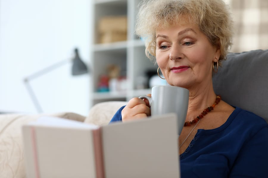 An older woman curled up on the couch reading a book and drinking tea.