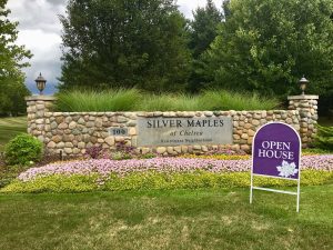 Large stone sign at the entrance of Silver Maples, surrounded by greenery and flowers, with an Open House sign in front